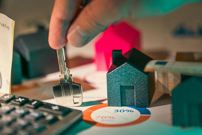 close up image of a hand placing a key on a table with house figures, calculator, and data reports about non-resident property mortgages
